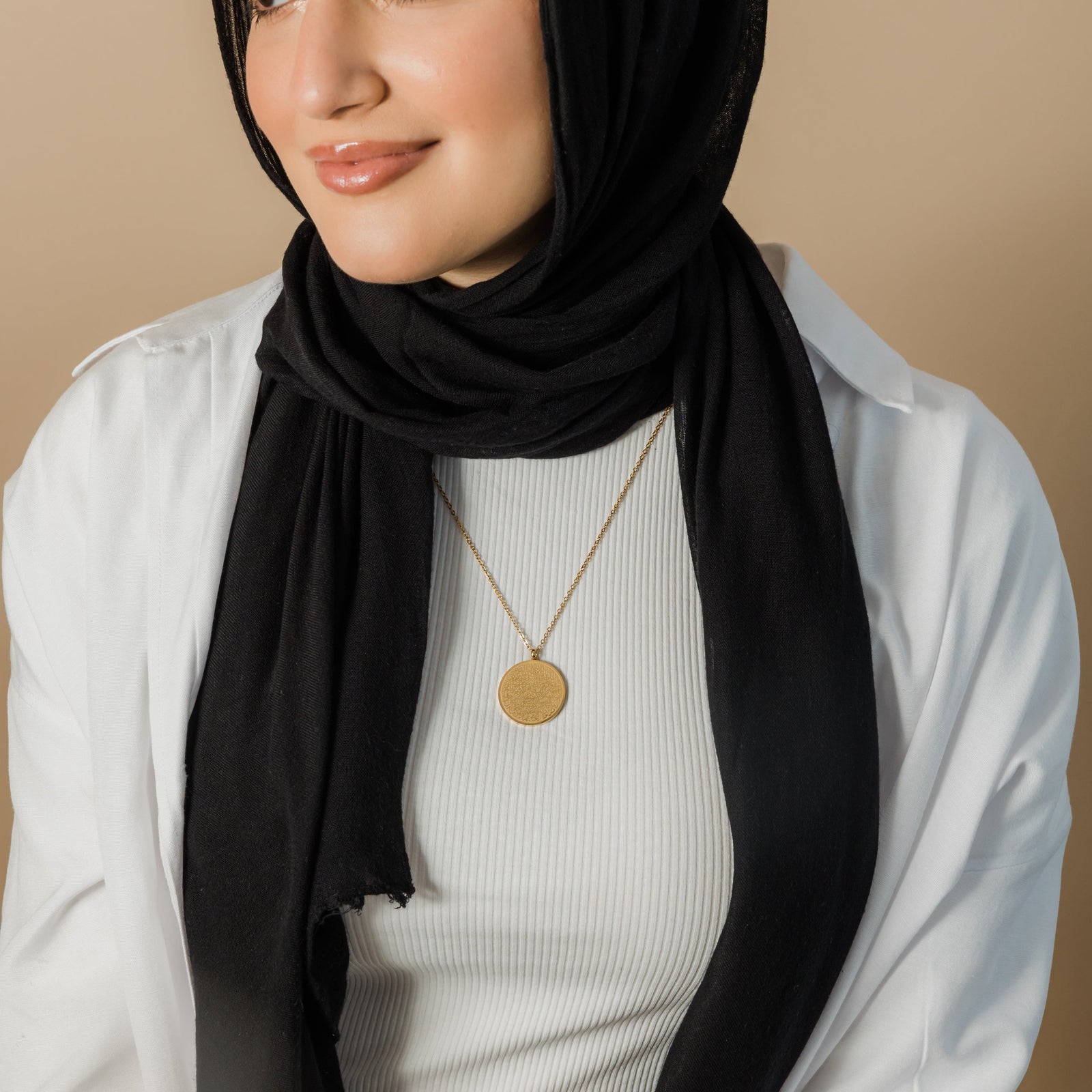 A person wearing a black hijab and a gold necklace with a round pendant inscribed with Arabic script is shown from the chest up. They are also wearing a white long-sleeved shirt over a white top. Given the inscription and the product name "Ayatul Kursi Necklace," it is highly likely that the script is from the Quran, a central religious text in Islam, suggesting a possible religious significance for the wearer. The person has light skin and is looking slightly to the right with a gentle smile.
