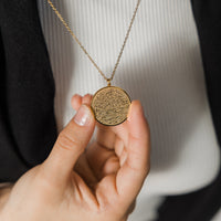 A hand holds a gold necklace with a round pendant inscribed with Arabic script. The chain is delicate and gold. The background is a white shirt and a black jacket. Given the inscription and the product name "Ayatul Kursi Necklace,"