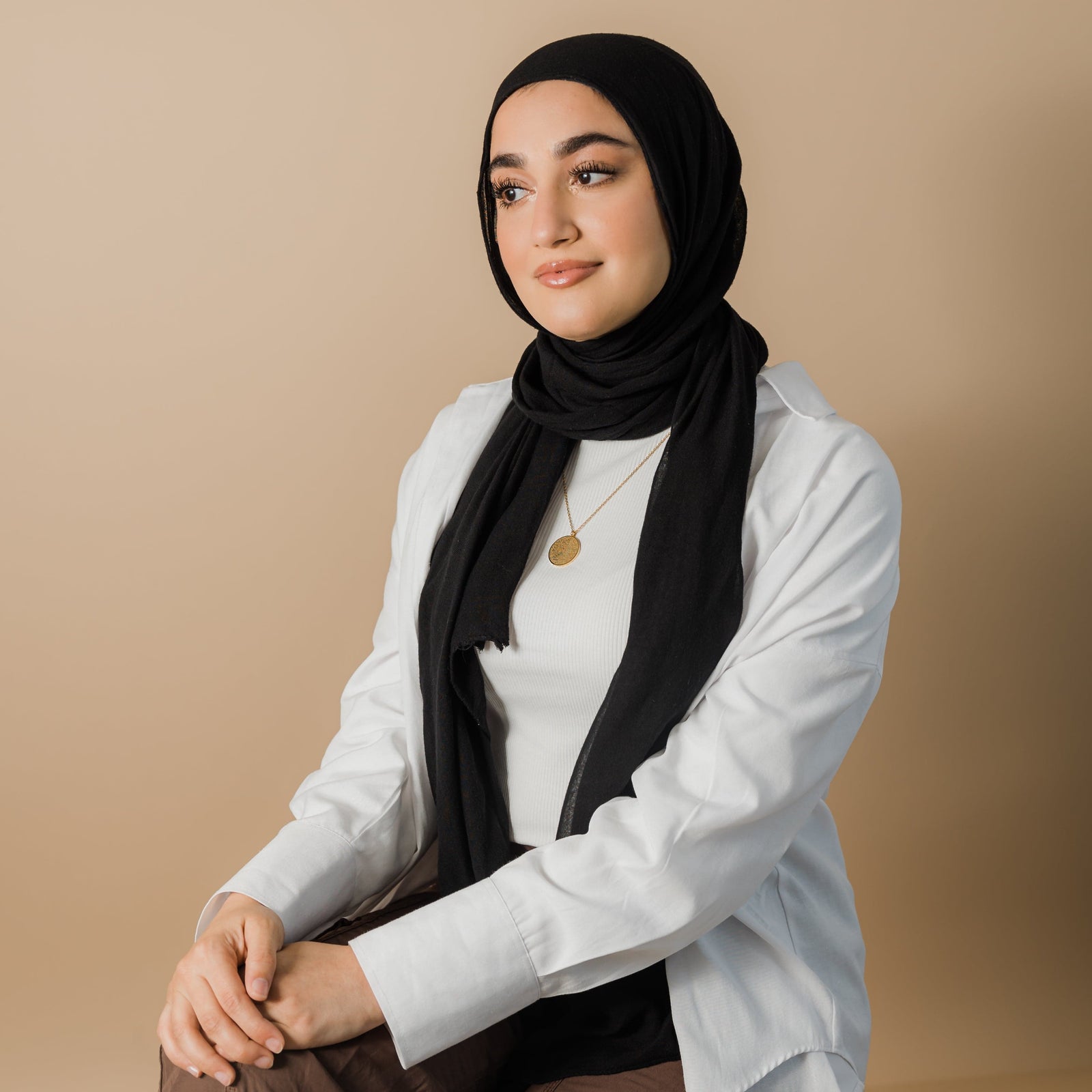 A person wearing a black hijab and a gold necklace with a round pendant inscribed with Arabic script is shown from the chest up. They are also wearing a white long-sleeved shirt over a white top. Given the inscription and the product name "Ayatul Kursi Necklace," it is highly likely that the script is from the Quran, a central religious text in Islam, suggesting a possible religious significance for the wearer. The person has light skin and is looking slightly to the right with a gentle smile.