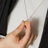 A delicate gold-plated necklace with a circular pendant. An Arabic letter is elegantly engraved in the center of the pendant. The necklace has a thin chain and is displayed on a light beige background. The specific Arabic letter is clearly visible within the circular frame of the pendant.