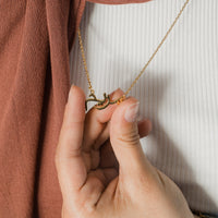 A close-up shows a hand gently holding a gold necklace with a pendant featuring a name written in elegant Arabic script. The necklace has a delicate gold chain. The background includes a white ribbed top and a portion of a rust-colored garment draped over the shoulder. The focus is on the intricate detail of the Arabic name pendant and the delicate nature of the jewelry.