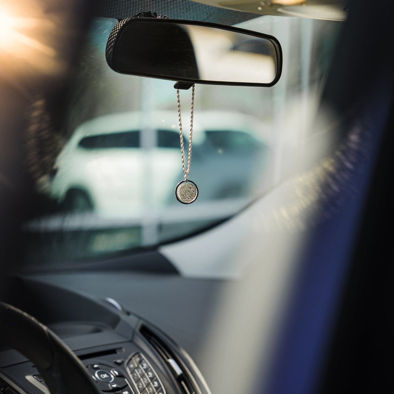 Silver car hanger with Shahada pendant hanging from a rearview mirror, photographed inside a car with blurred background.