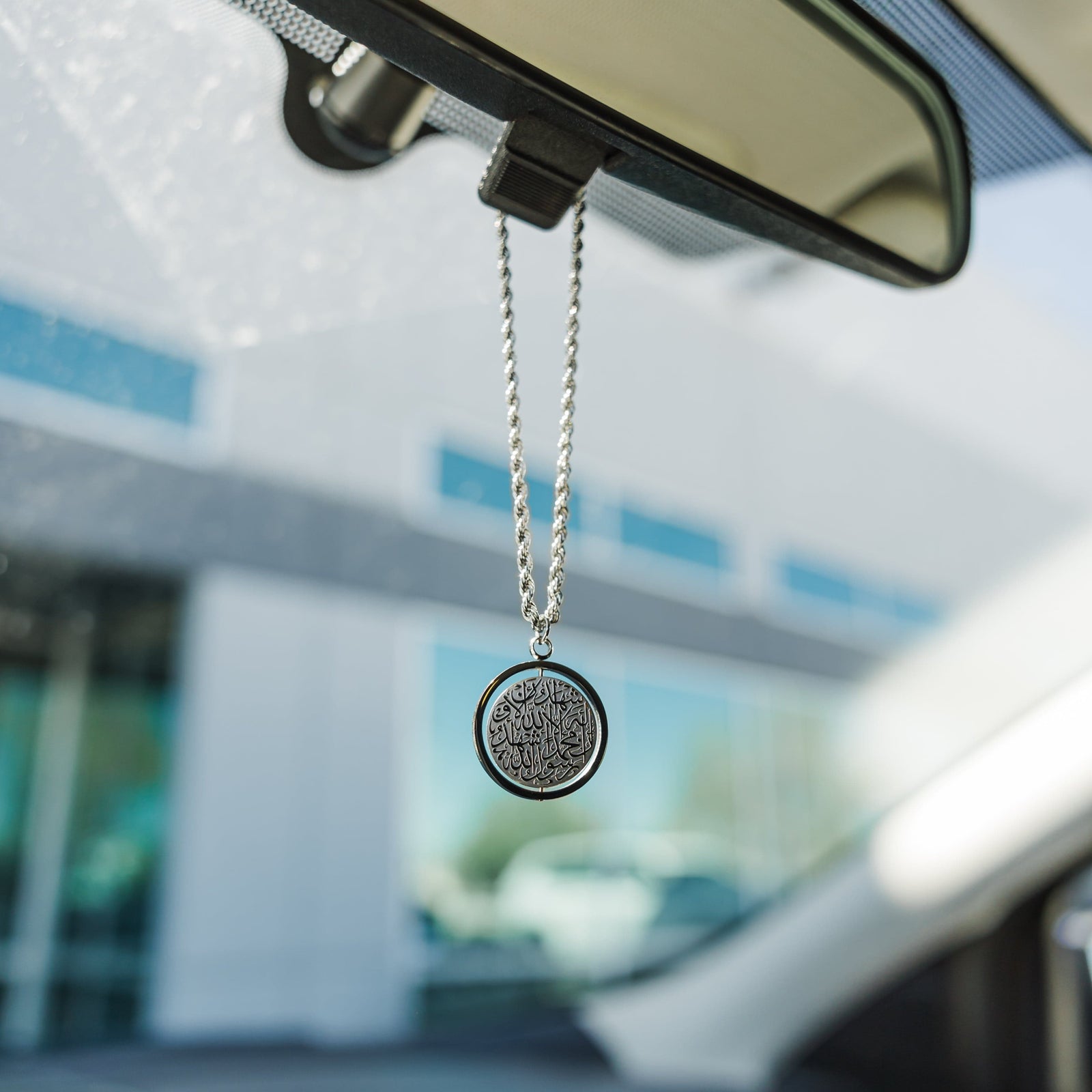 Silver rope chain car hanger with a circular pendant engraved in Arabic calligraphy of the Shahada, hanging from a rearview mirror with blurred outdoor background.