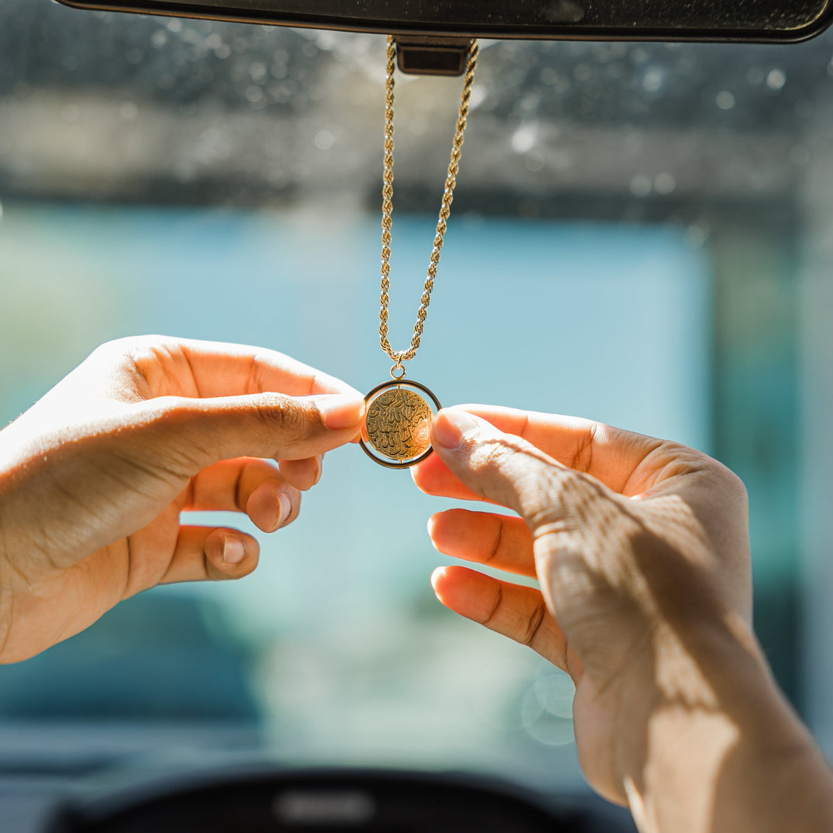 Hands holding and adjusting a gold car hanger with a circular Shahada pendant, hanging from a rearview mirror inside a car.