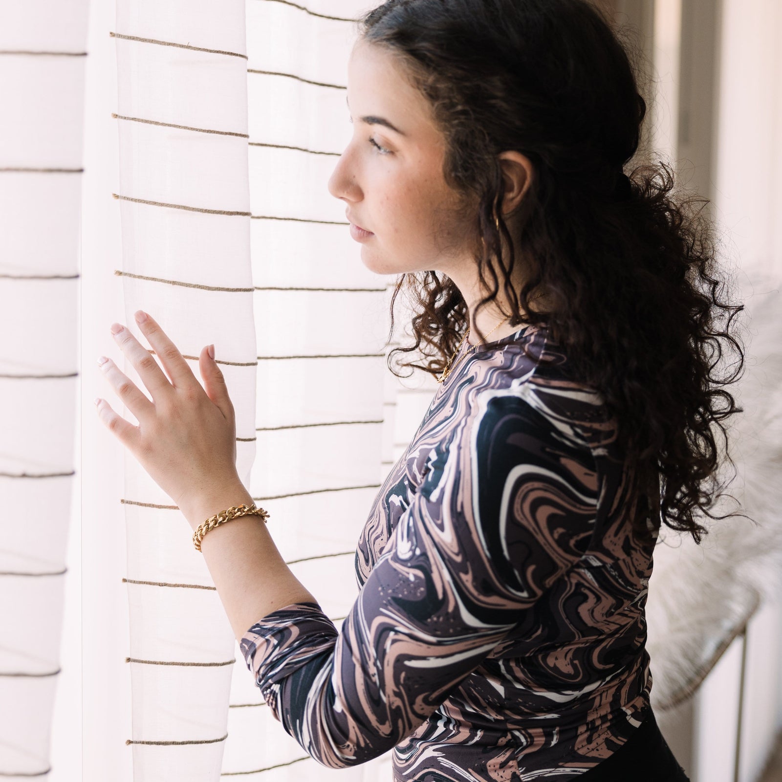 Photo of the model looking out the window. The model is wearing the gold Chunky Cuban Chain Bracelet on their left hand.