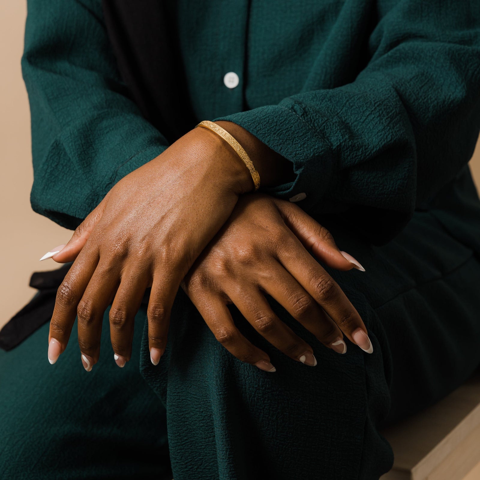 Close up of the model's hands resting over her lap. The model is wearing the gold Ayatul Kursi Cuff.