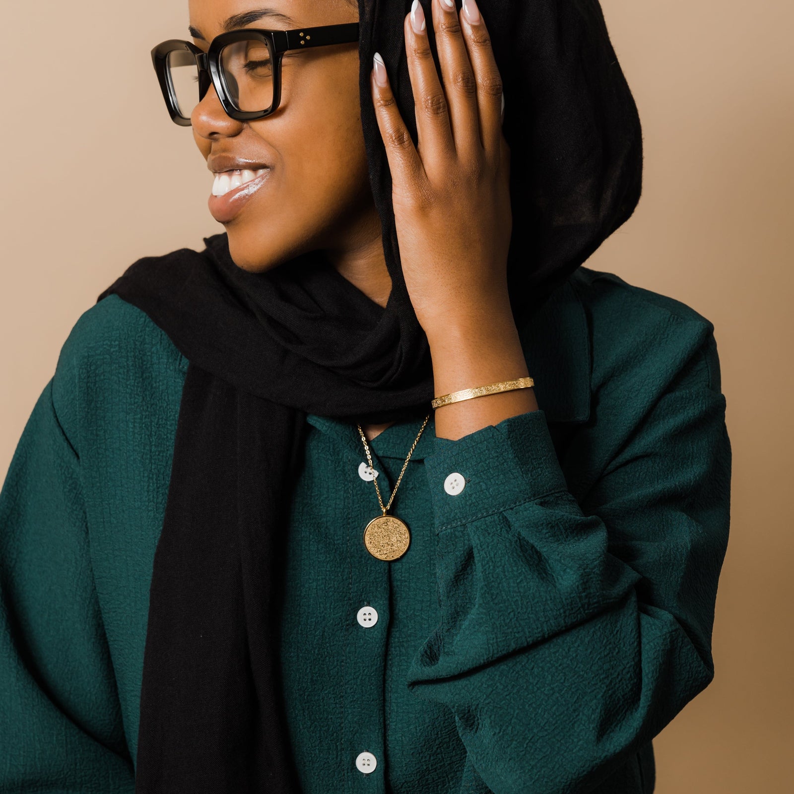 Photo of the model looking away from the camera and touching their black hijab with their left hand. The model is wearing the gold Ayatul Kursi Necklace and Ayatul Kursi Cuff.