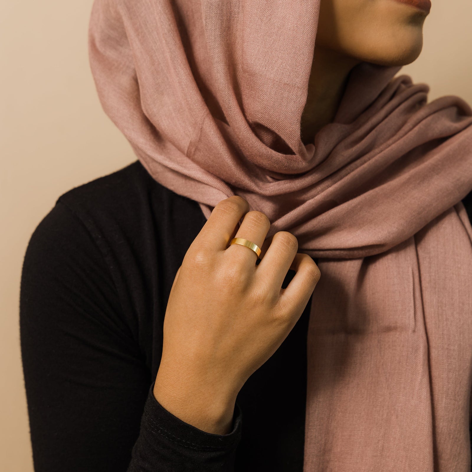 Side view of a woman in a dusty rose hijab wearing a single flat gold band ring on her finger.