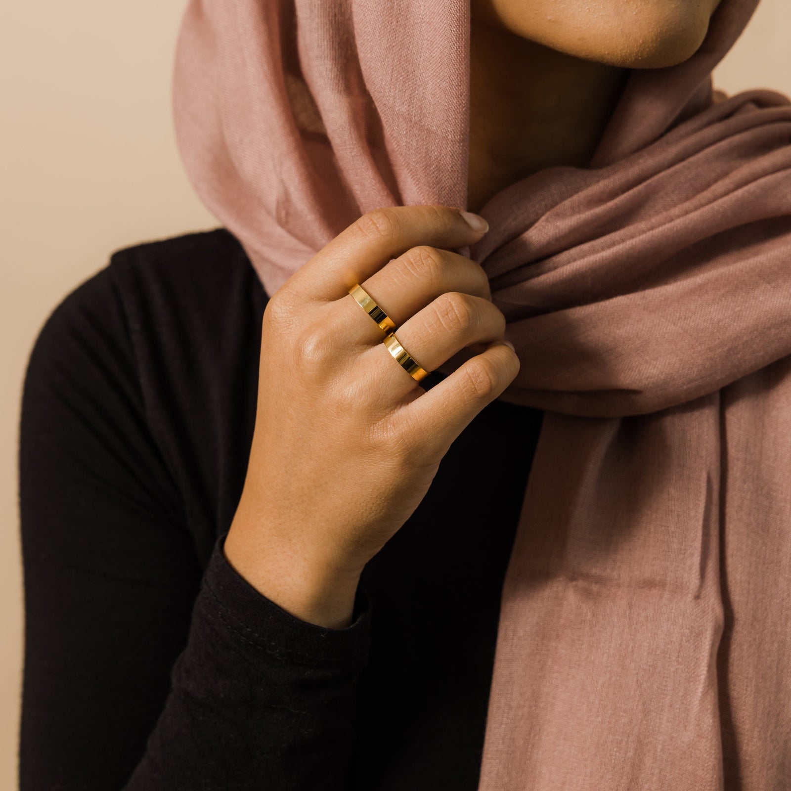 Woman wearing two gold rings, holding dusty rose hijab near her face.