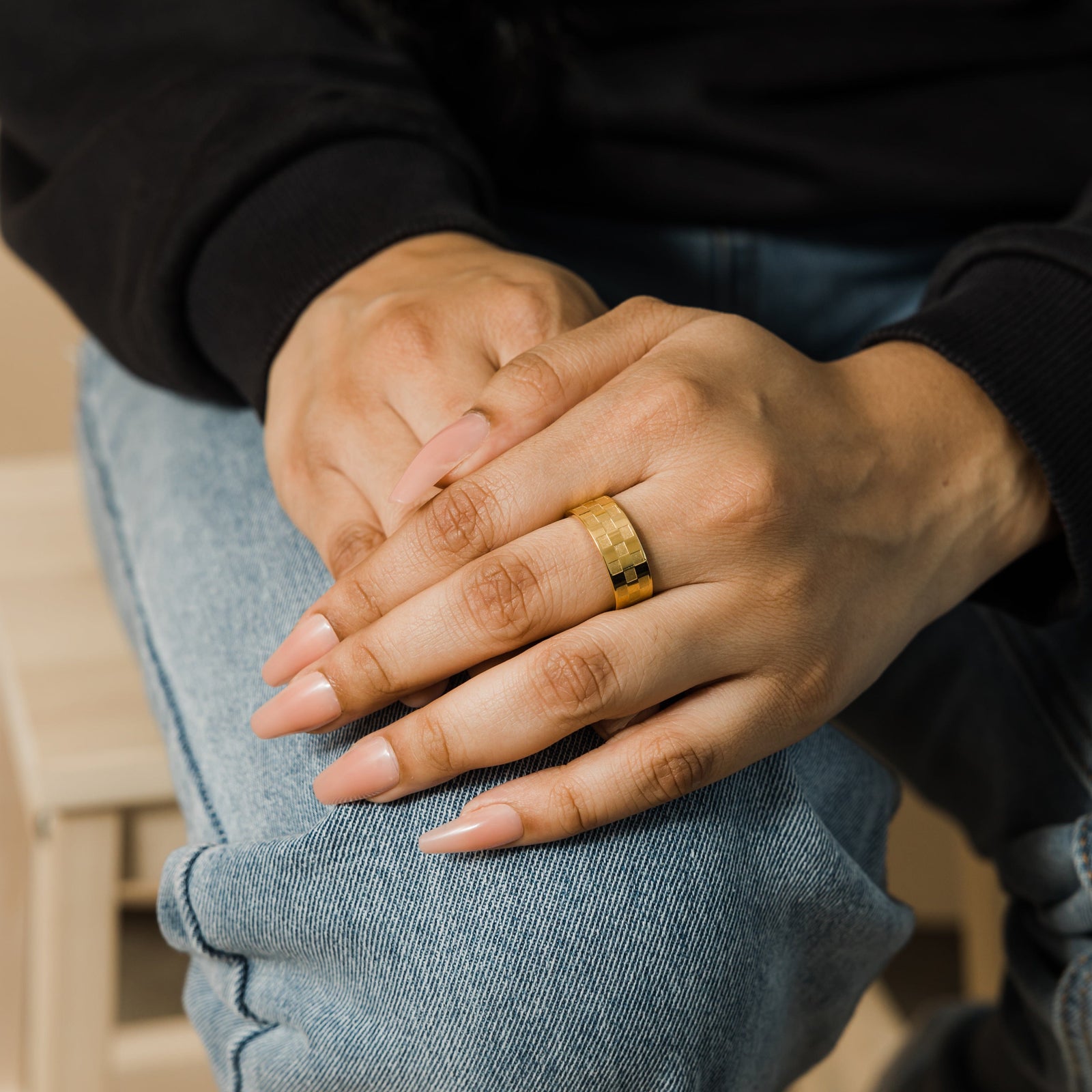 Gold Queen’s Ring with bold checkered design worn on middle finger, hands resting together casually over light denim jeans.