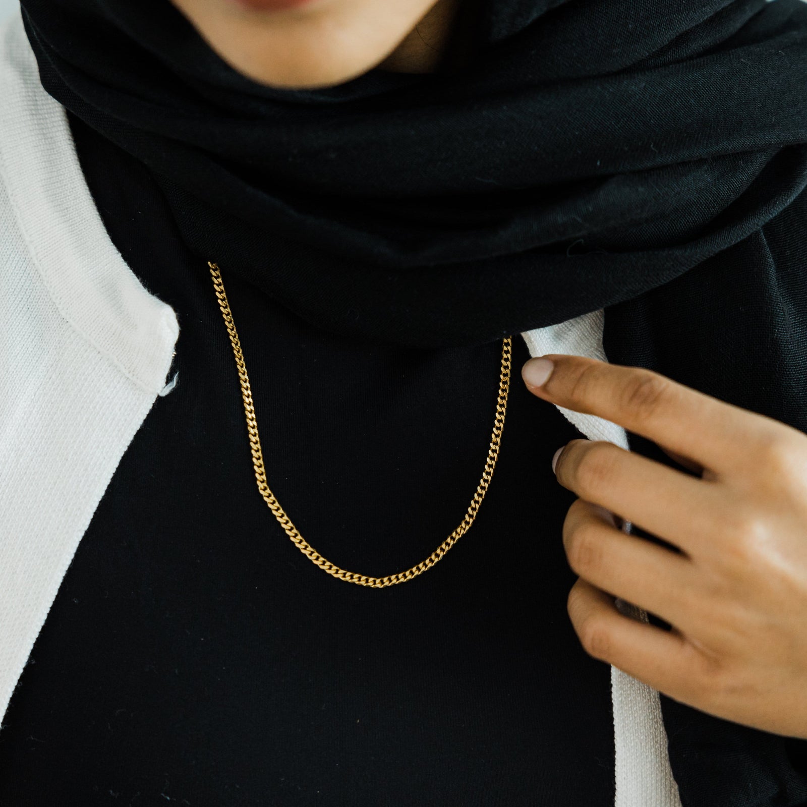 A woman wearing a Cuban chain necklace over a black hijab and top, with fingers placed on the chest.