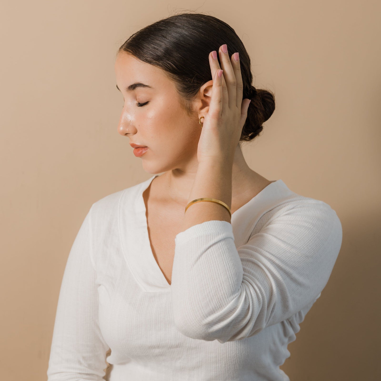 Photo of the model with their head turned to the side with their eyes closed. The model is touching their hair with their left fingers to show the gold Essential Bangle they are wearing on their left hand.