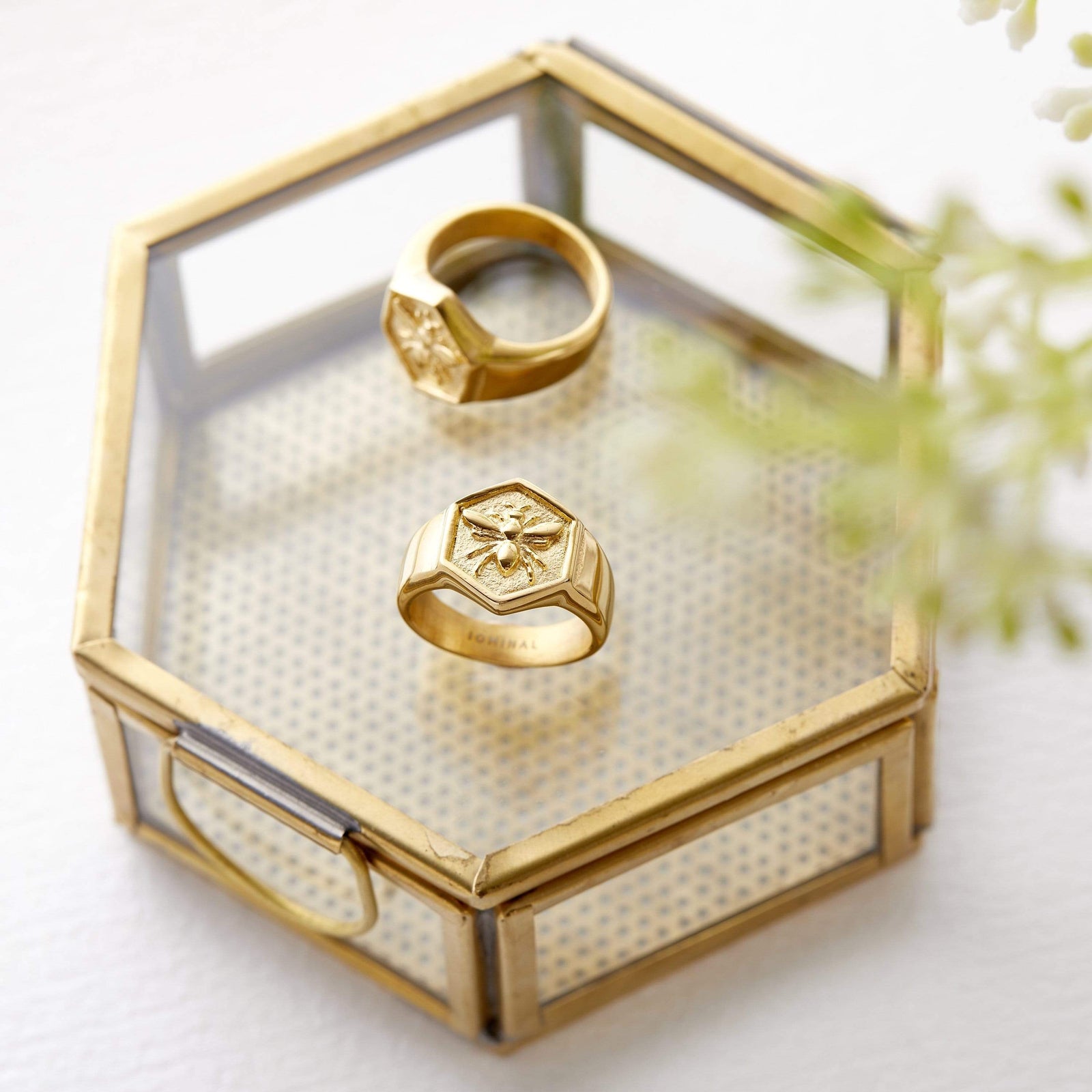 Two gold Nahla Rings with bee designs placed on a glass hexagon jewelry box with gold edges.