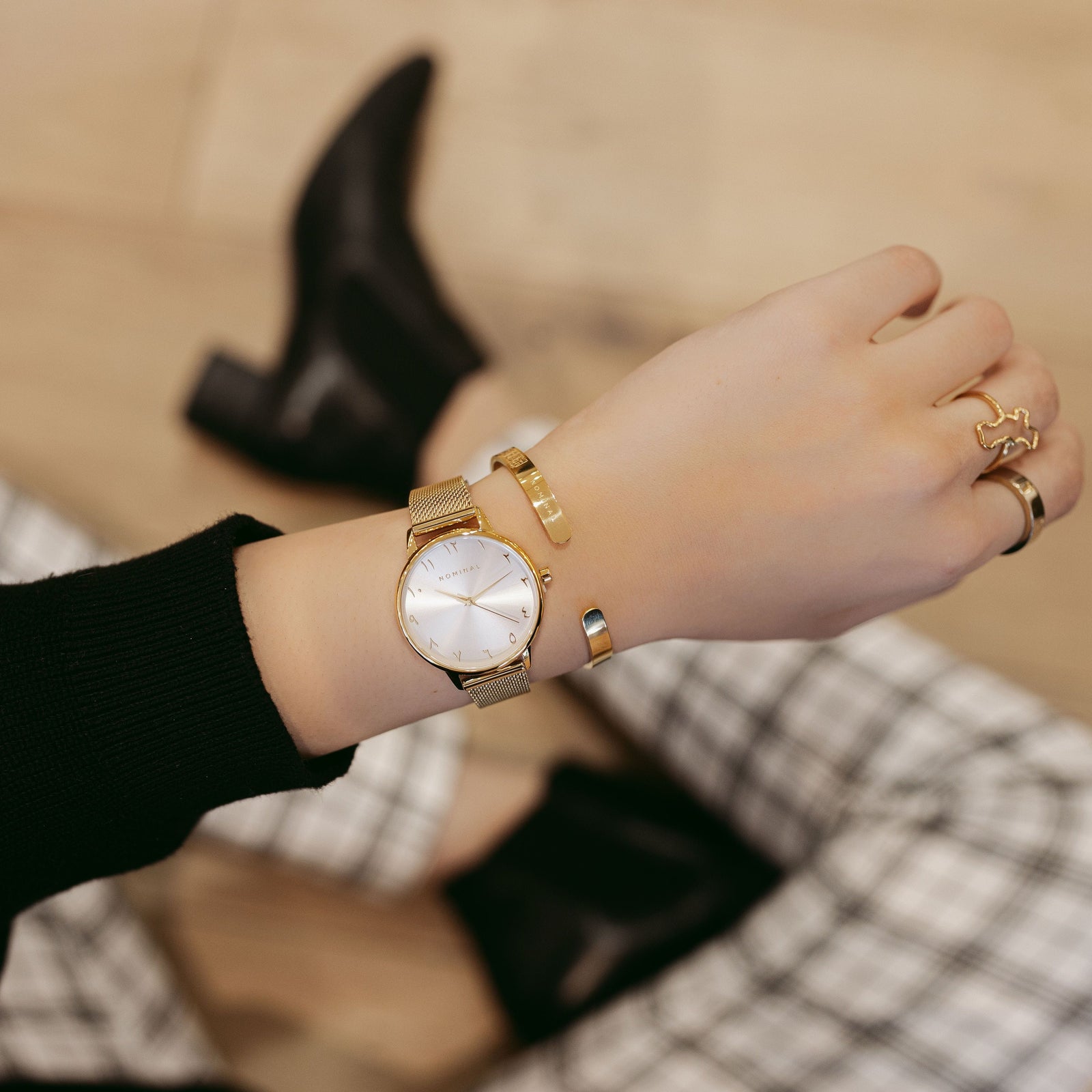 Gold mesh strap watch with white background and gold details, with minimalist Arabic numerals worn on the wrist of a women wearing black and white striped pants, black boots and a black sweater and matching gold cuff and rings. 
