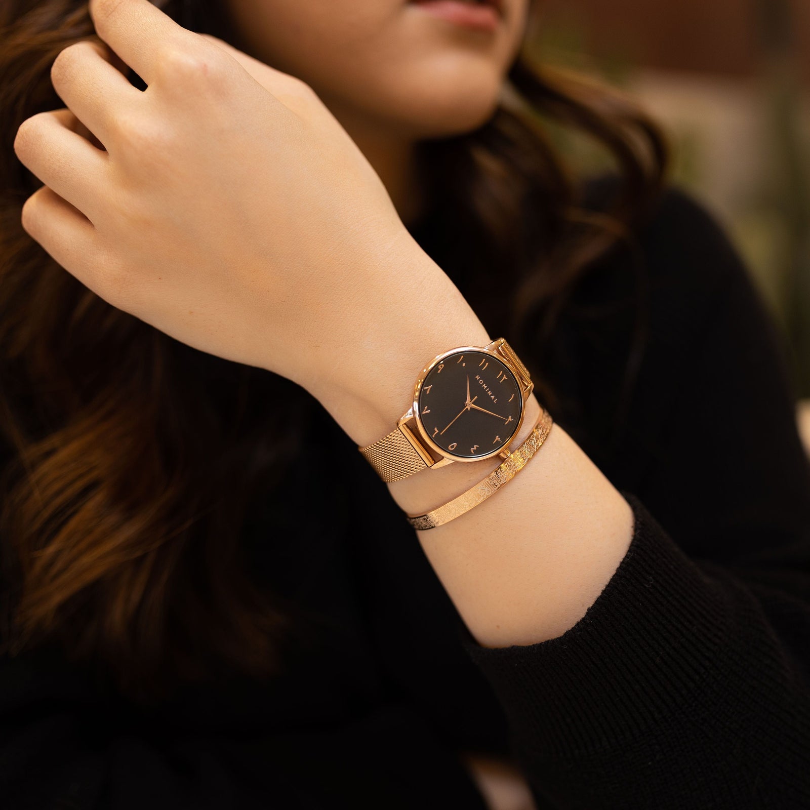 Close-up of a gold mesh strap watch with minimalist Arabic numerals worn on the wrist of a woman against a black sweater. The woman is also wearing a gold cuff. 