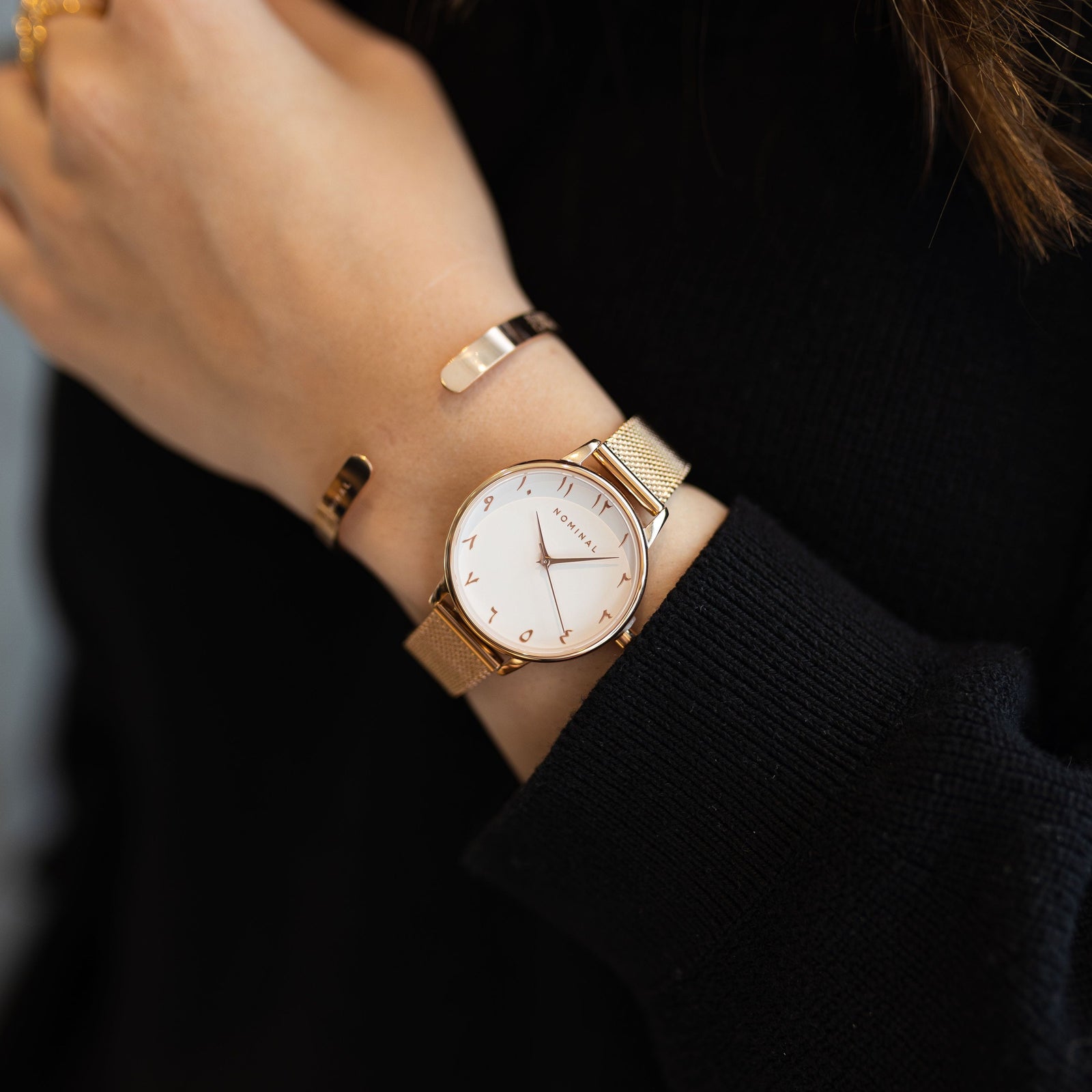 Close-up of a rose gold mesh strap watch with Arabic numerals, worn on the wrist of a woman against her black sweater.