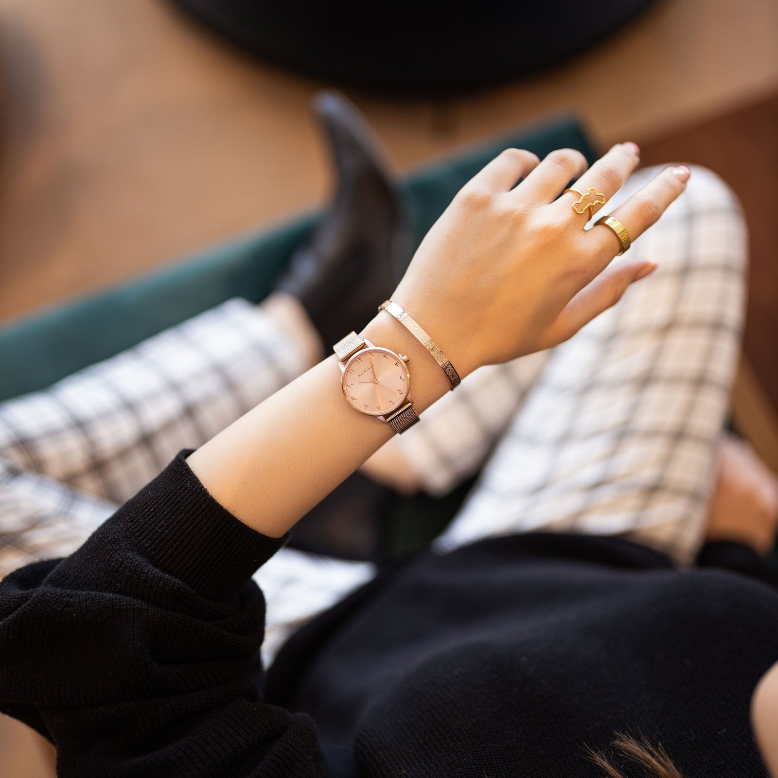 Close-up of a rose gold mesh strap watch with Arabic numerals, worn on the wrist of a woman over her white and black striped pants who is sitting on a chair.