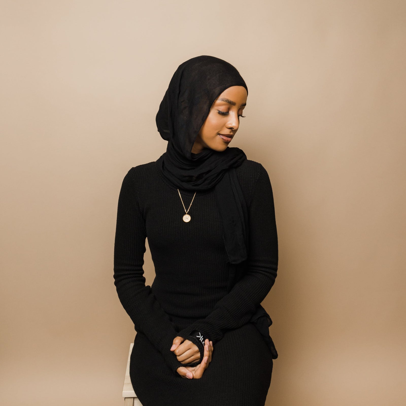 Photo of the model looking down with their hands clasped on their lap. The model is wearing the gold Patience Token Necklace.