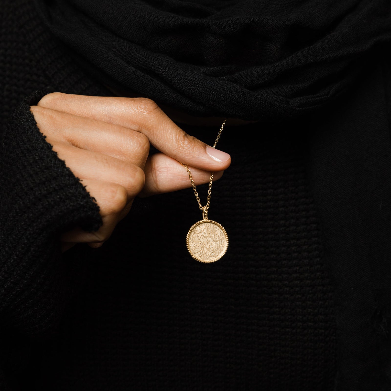 close up of the gold Patience Token Necklace as the model lifts the chain to show the design of the pendant.