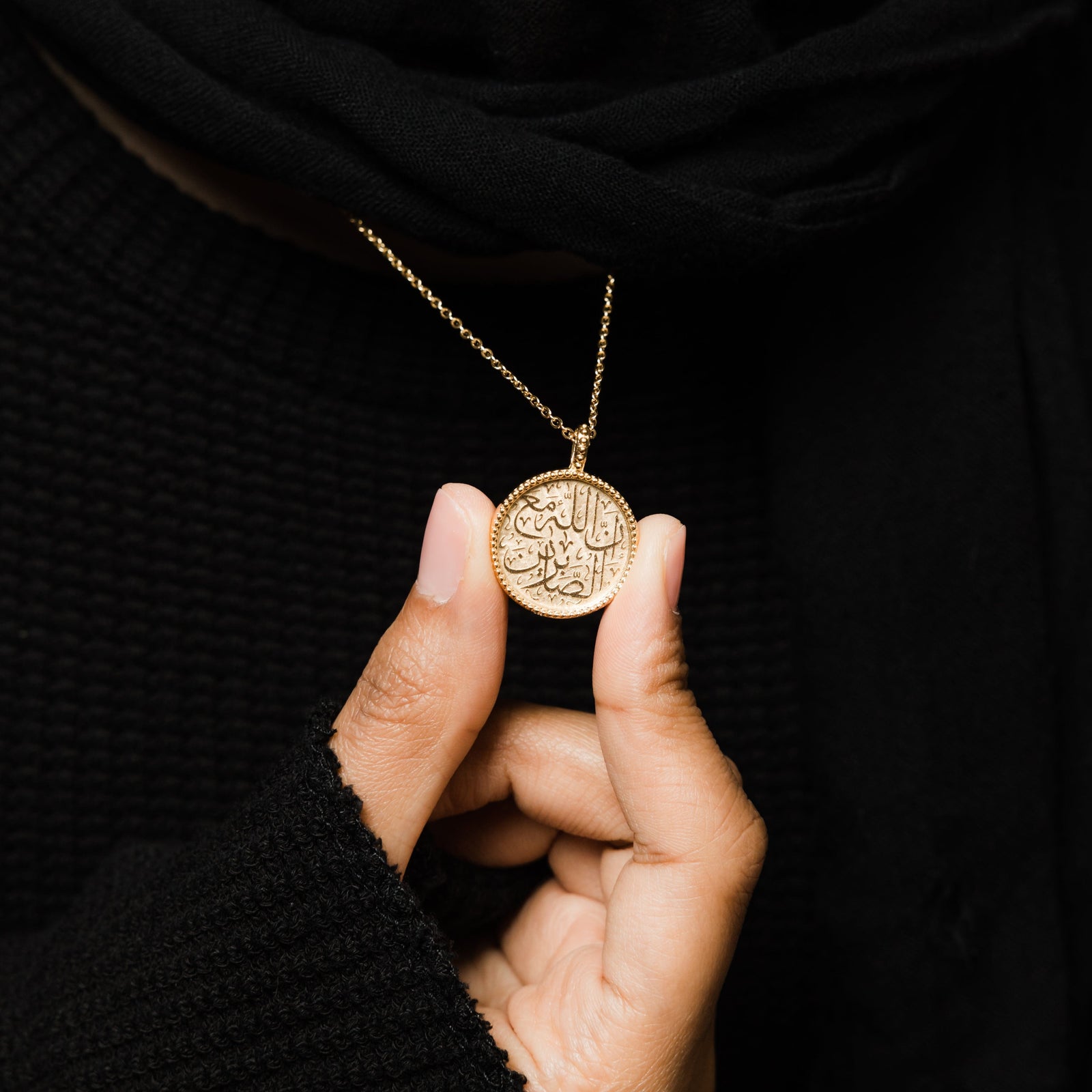 Close up of the gold Patience Token Necklace against the black fabric of the model's clothing. The model also holds the pendant between their fingers to showcase its design.