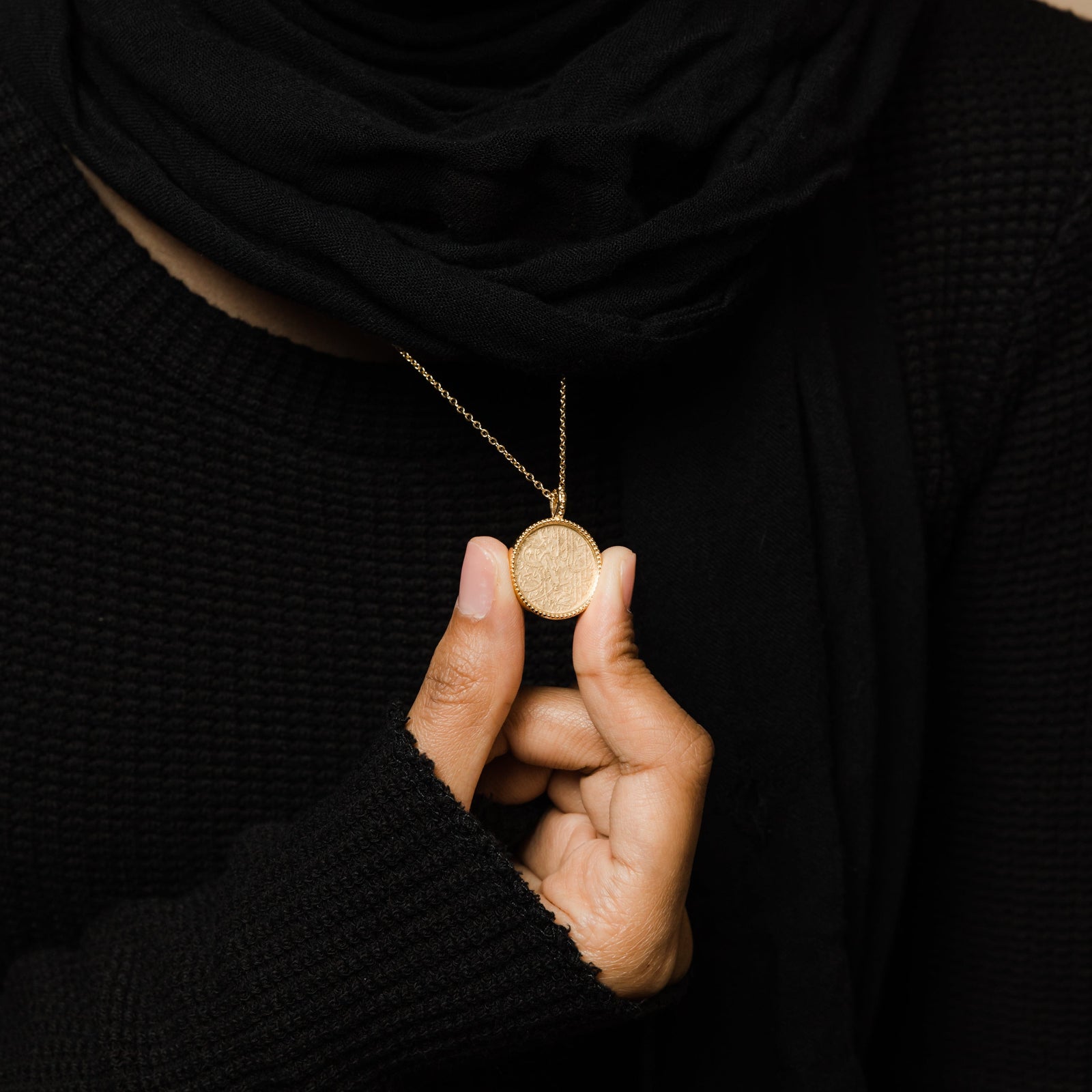 Close up of the gold Patience Token Necklace as the model holds the pendant to show its design.