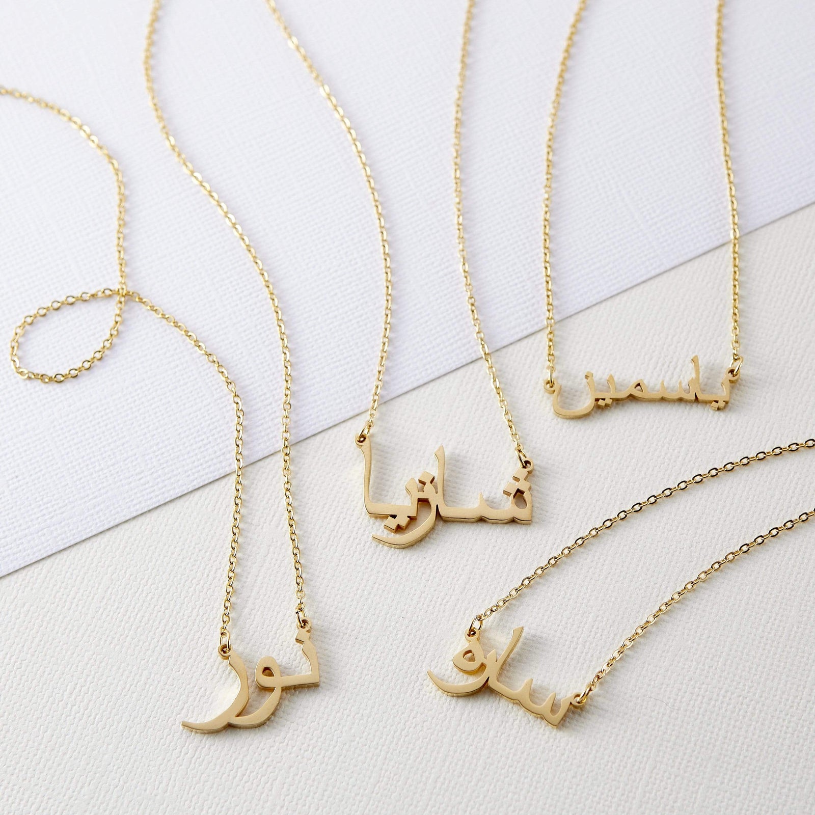 Four gold necklaces with pendants featuring different names written in Arabic script are arranged on a white rectangular tray. Each necklace has a delicate gold chain. The names on the pendants are distinct in their calligraphic style. The tray rests on a textured, light-colored surface.