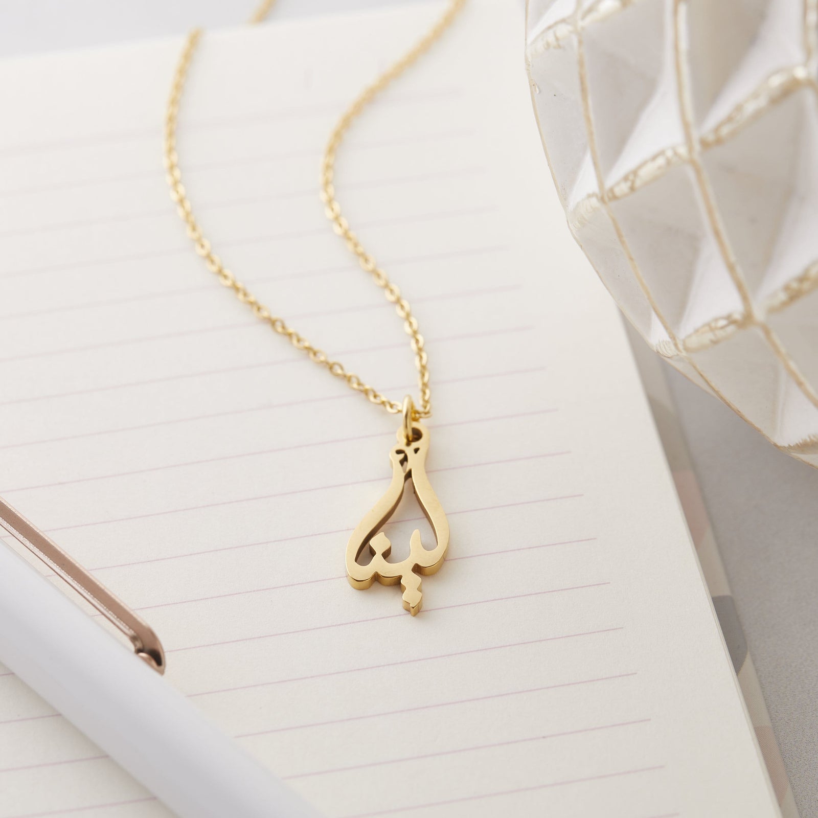 Close up of the gold Ready Calligraphy Name Necklace - Lena against a light background.