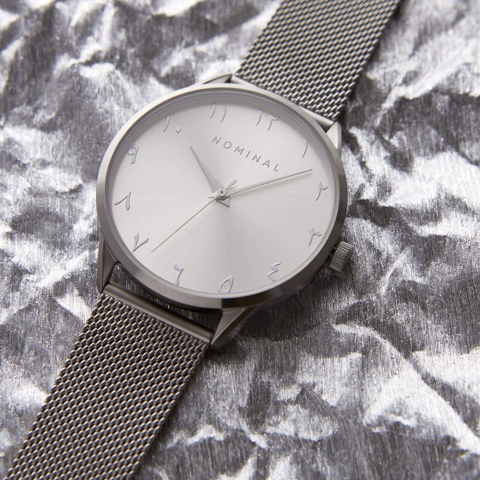 Photo of a watch with silver case and silver mesh strap. The round white sunburst dial features silver numbers. Nominal Logo, and pin.