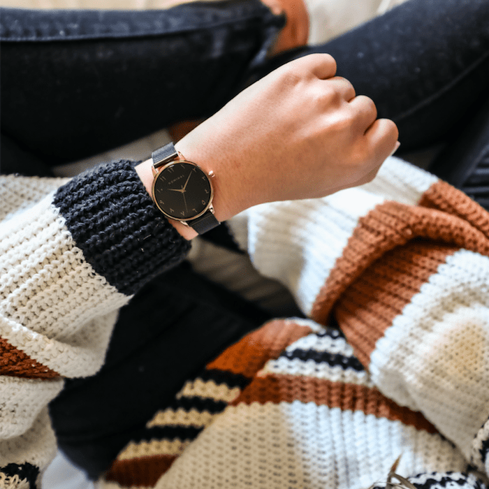 Close-up of a black watch with rose gold r details, with minimalist Arabic numerals worn on the wrist of a woman sitting down wearing jeans and a white, brown and black striped sweater.