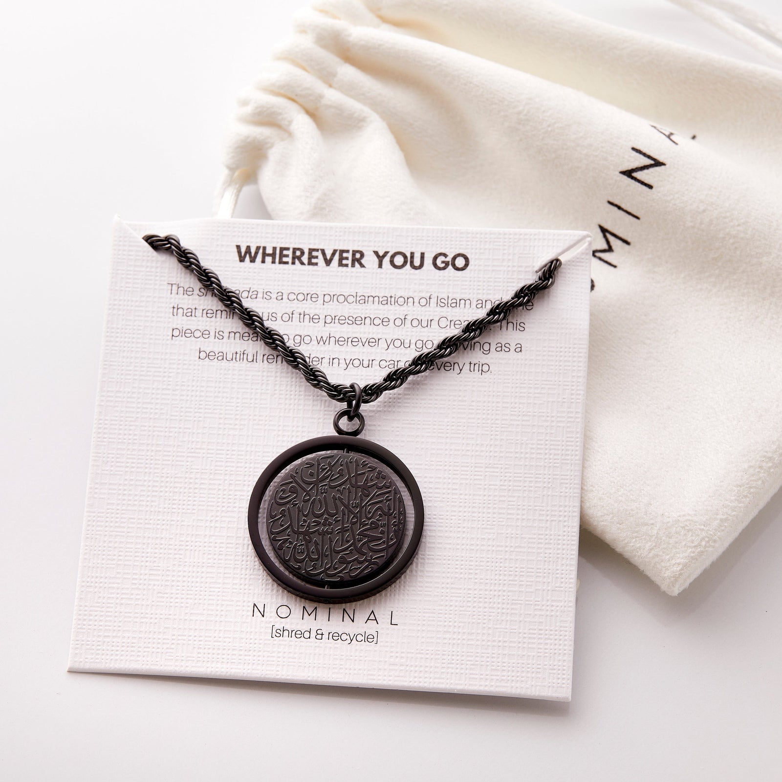 Black  Shahada car hanger displayed on a Nominal card that reads “Wherever You Go” with descriptive text underneath, paired with a white drawstring pouch in the background. The circular pendant is engraved in Arabic calligraphy of the Shahada.