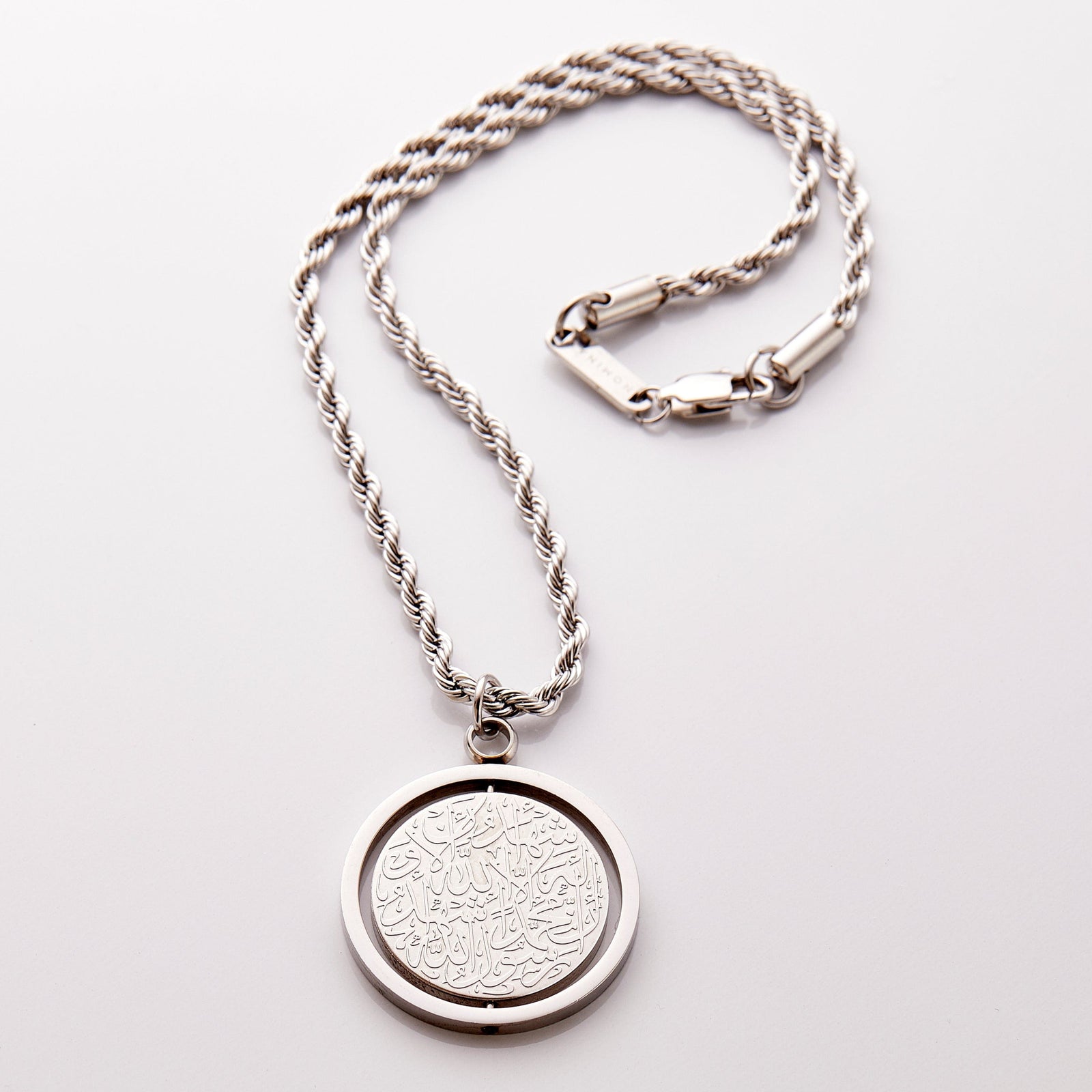 Silver car hanger with a circular pendant engraved in Arabic calligraphy of the Shahada, styled flat against a neutral background.