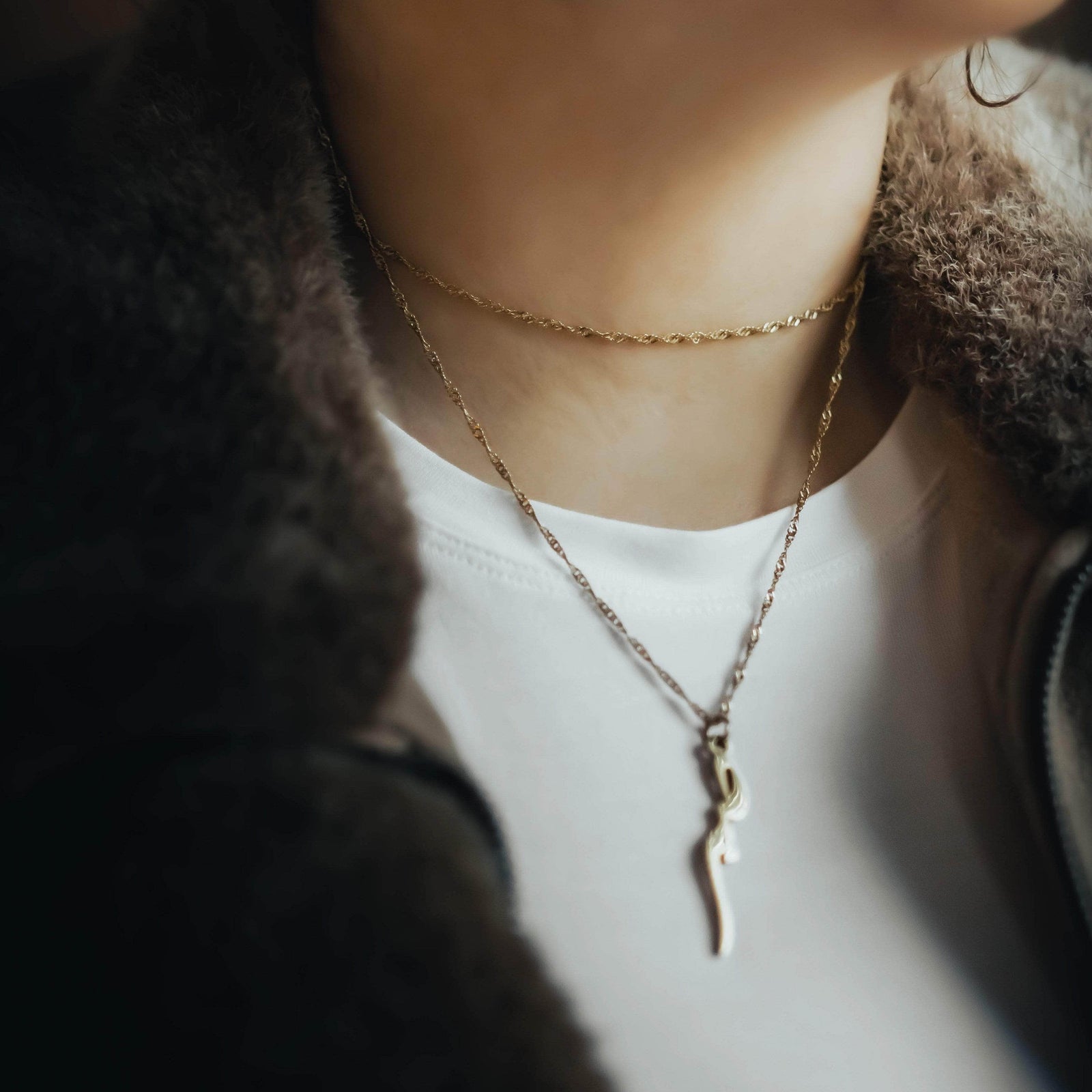 Close-up of a gold twisted chain choker layered with a longer pendant necklace, styled over a white top and brown jacket.