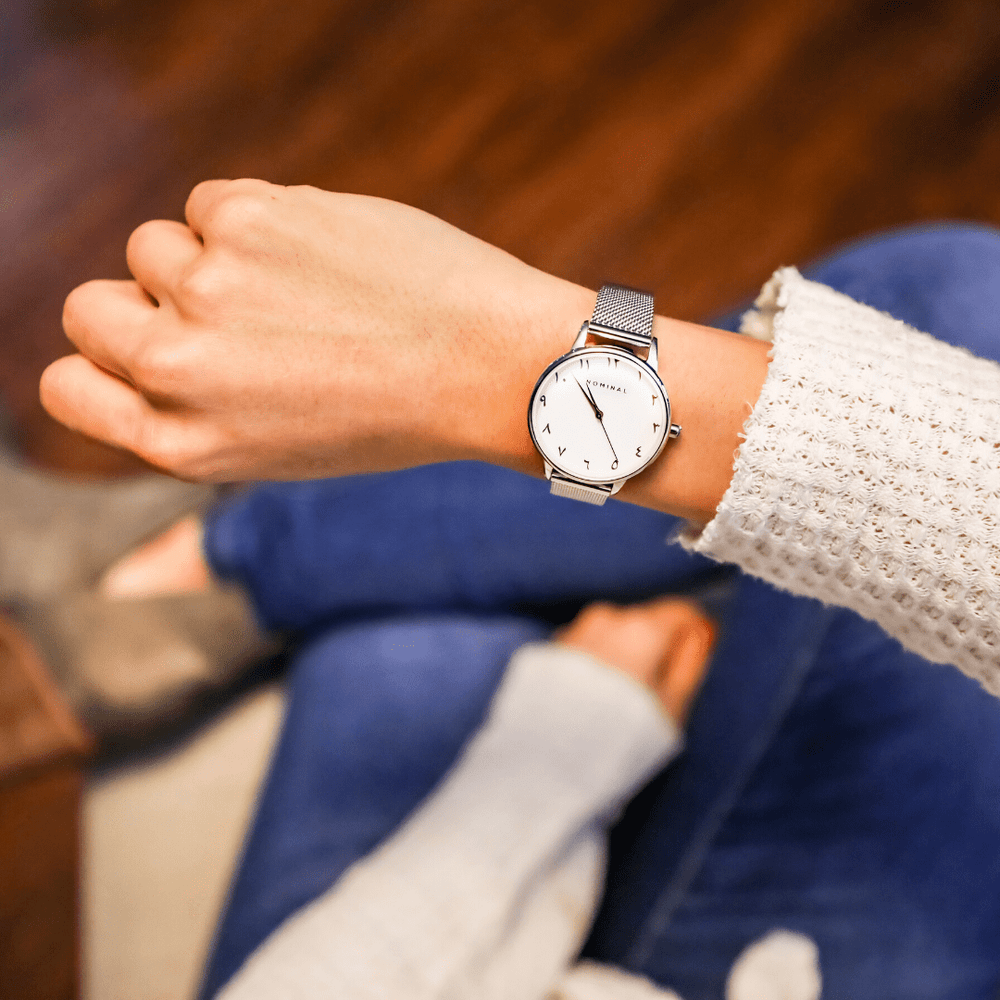 Close-up of a silver mesh strap watch with white background and silver details, with minimalist Arabic numerals worn on the wrist of a woman sitting down wearing denim jeans and a white sweater. 