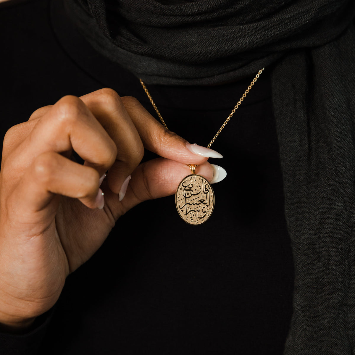 Close up of the gold "Verily, with Hardship Comes Ease" Oval Necklace as the model holding the pendant.