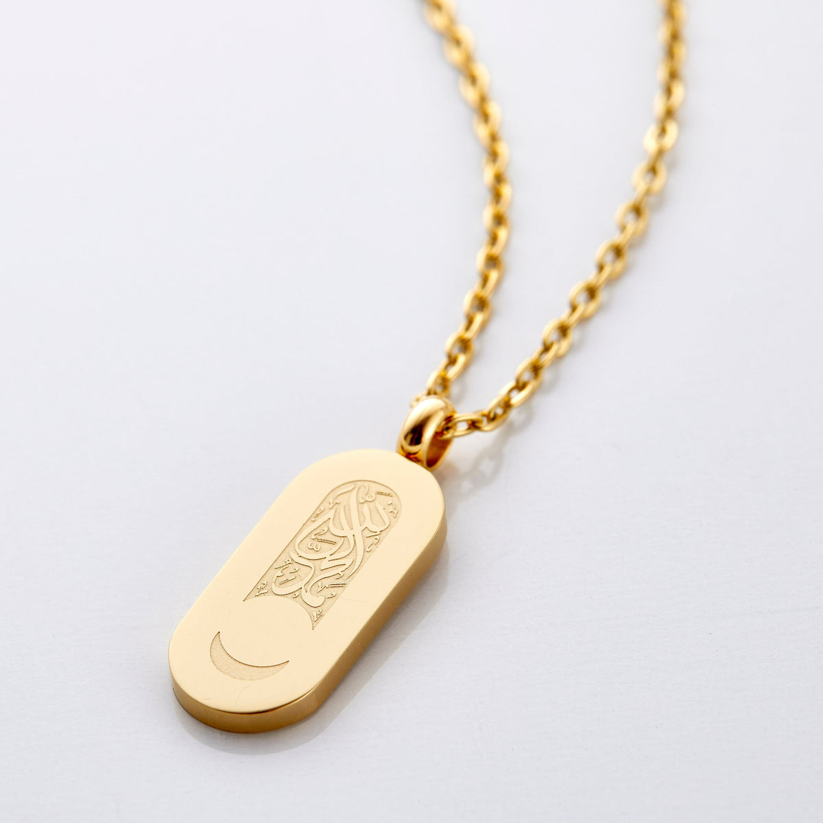 An 18k gold plated necklace that has a pill-shaped pendant that contains a blank space below the Arabic calligraphy to leave a remaining crescent at the bottom of the tag.