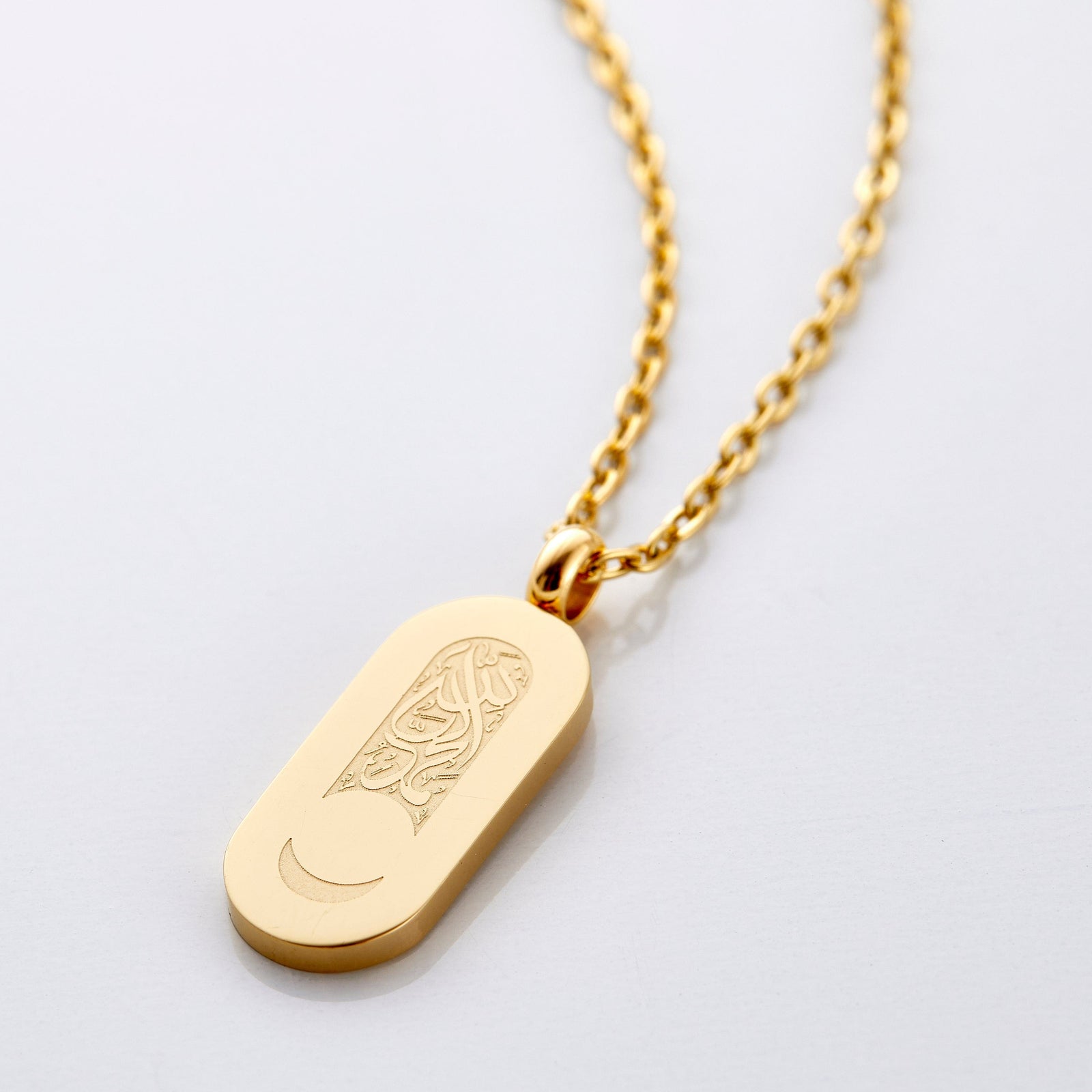 An 18k gold plated necklace that has a pill-shaped pendant that contains a blank space below the Arabic calligraphy to leave a remaining crescent at the bottom of the tag.