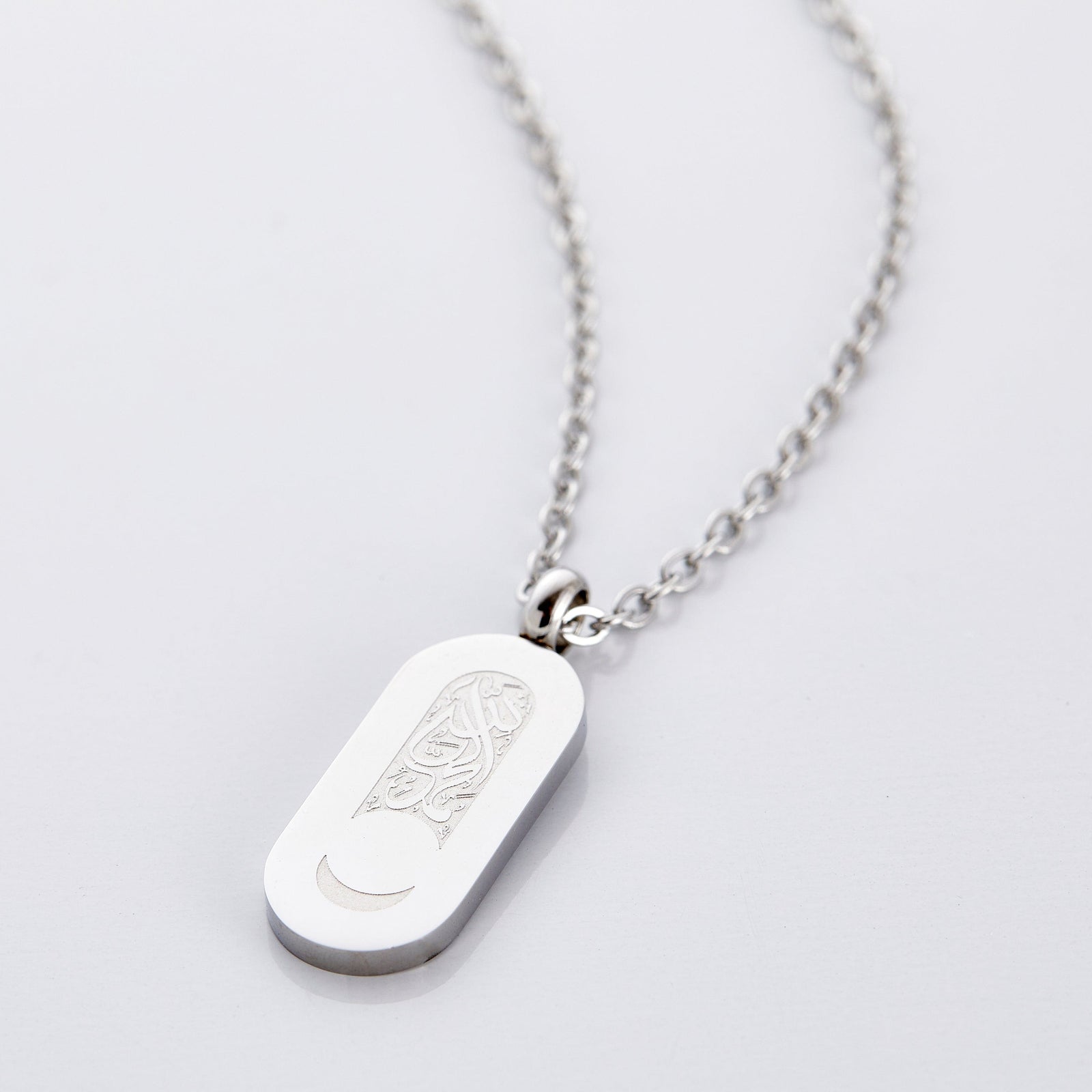 A silver necklace that has a pill-shaped pendant that contains a blank space below the Arabic calligraphy to leave a remaining crescent at the bottom of the tag.