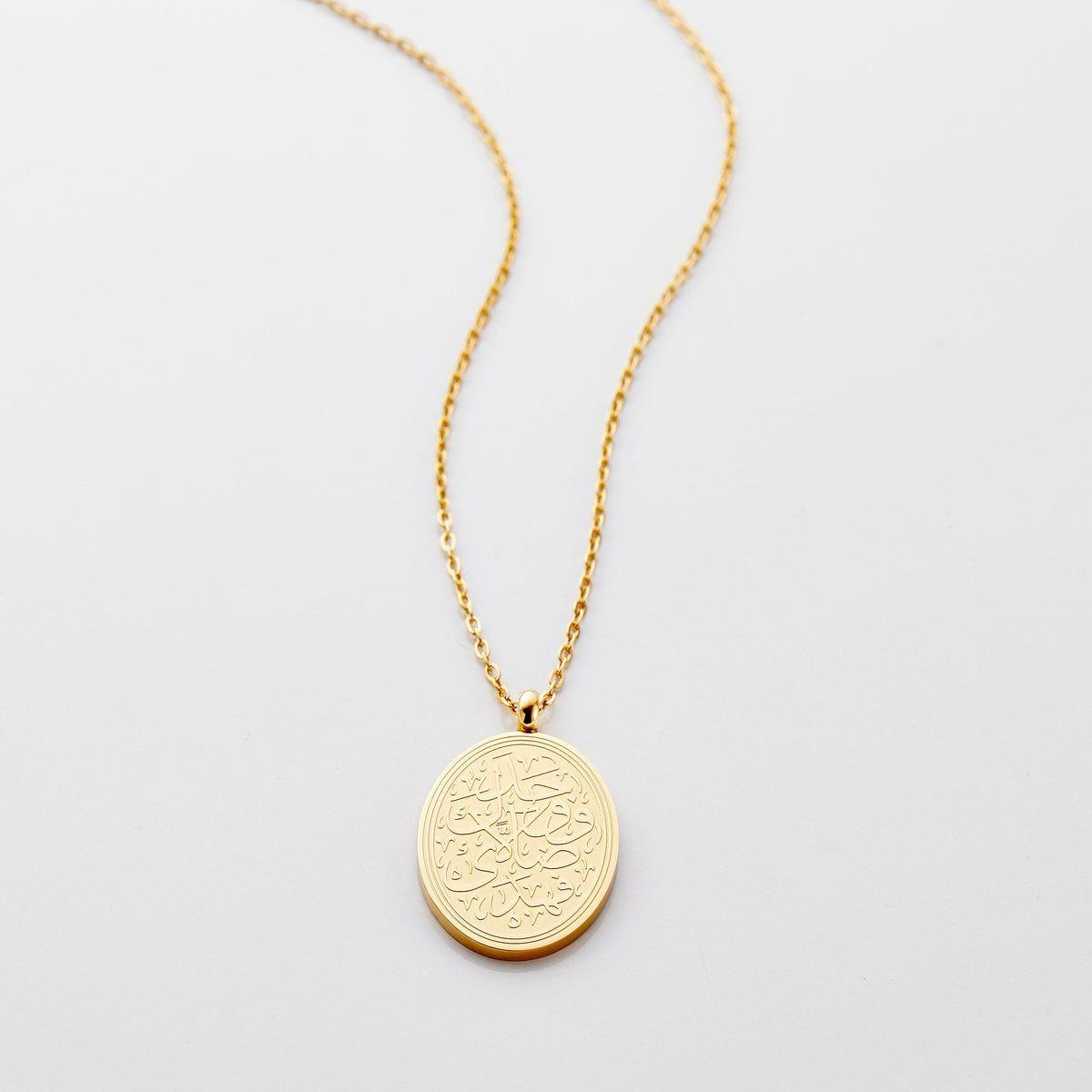 Flat lay of an 18k gold plated earrings that features a round pendant. This pendant is surrounded by ridged lines along the edges to accentuate the beautiful swirling calligraphy in the center. 