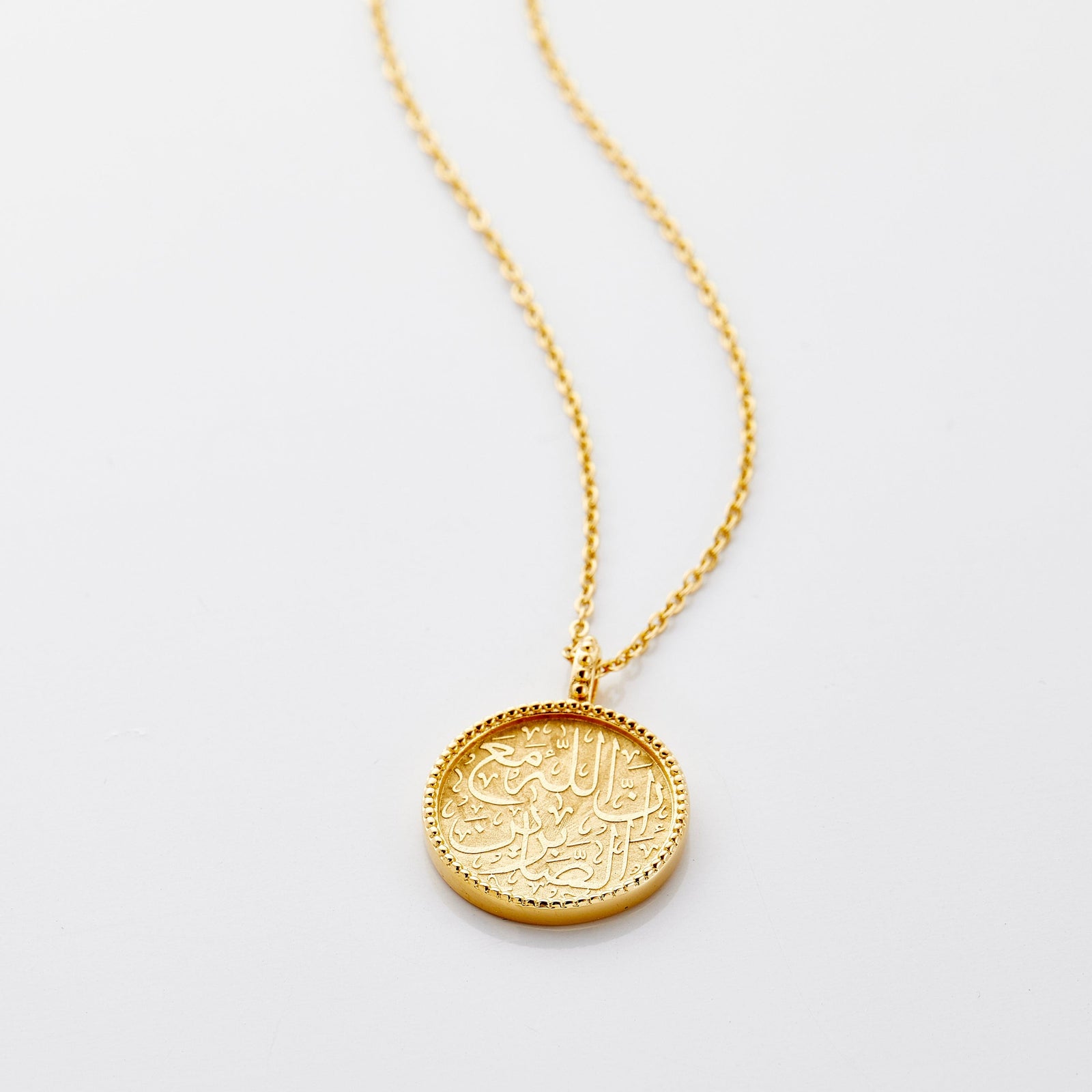 Photo of an 18k gold plated necklace with a round pendant that features Arabic calligraphy engraving and  vintage fading and ridged edges around the pendant.