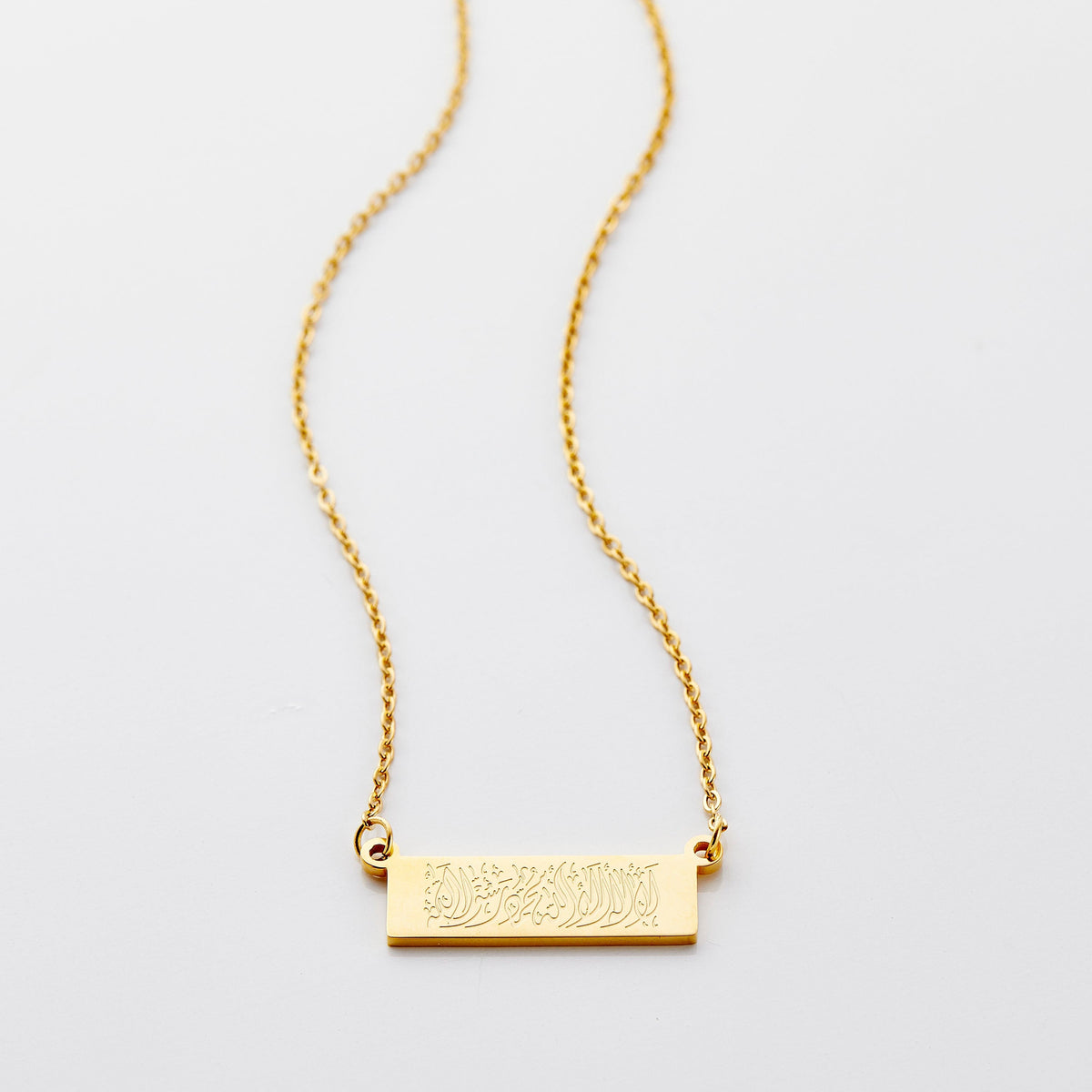 Flat lay of an 18k gold plated necklace that features a bar pendant with Arabic calligraphy engraving.