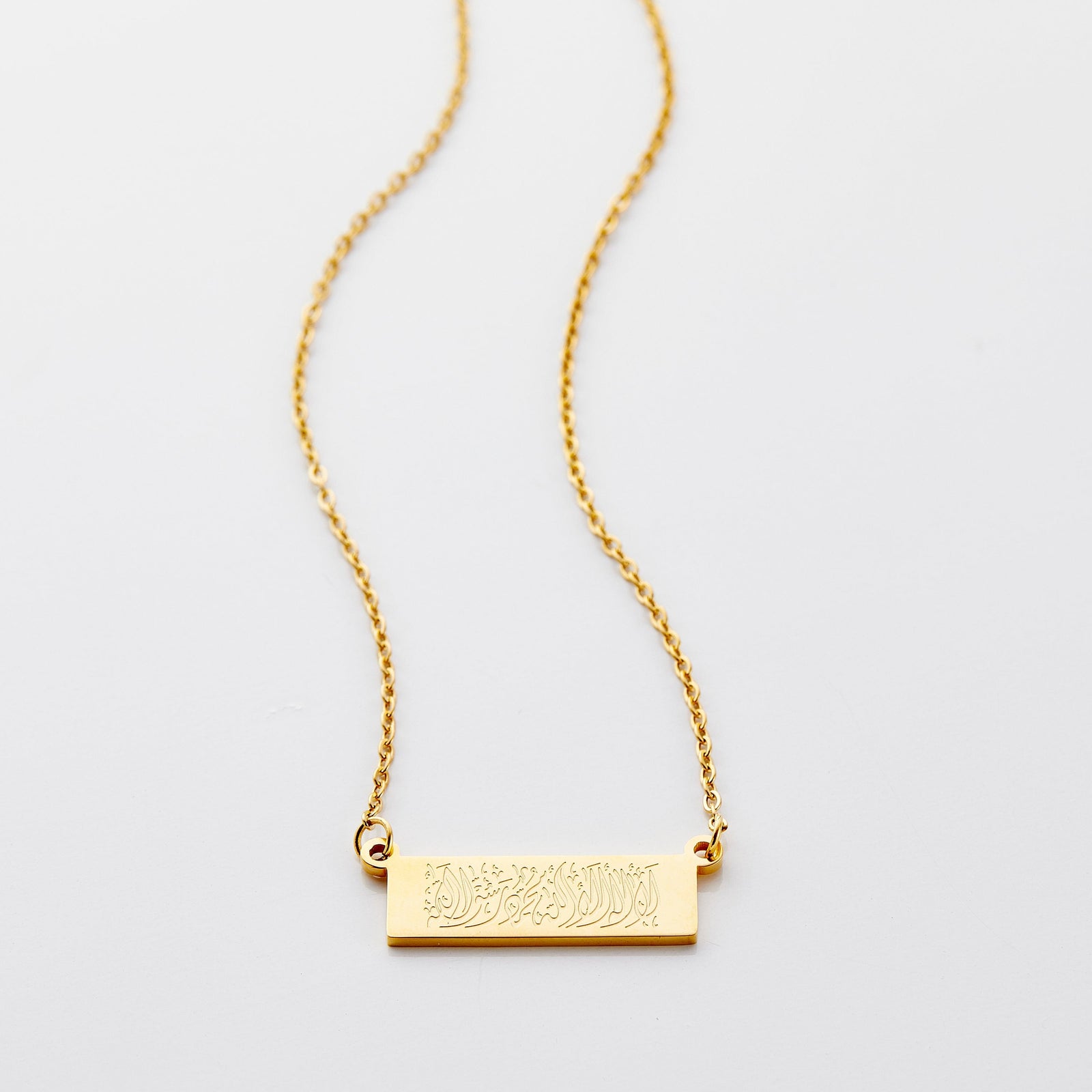 Flat lay of an 18k gold plated necklace that features a bar pendant with Arabic calligraphy engraving.