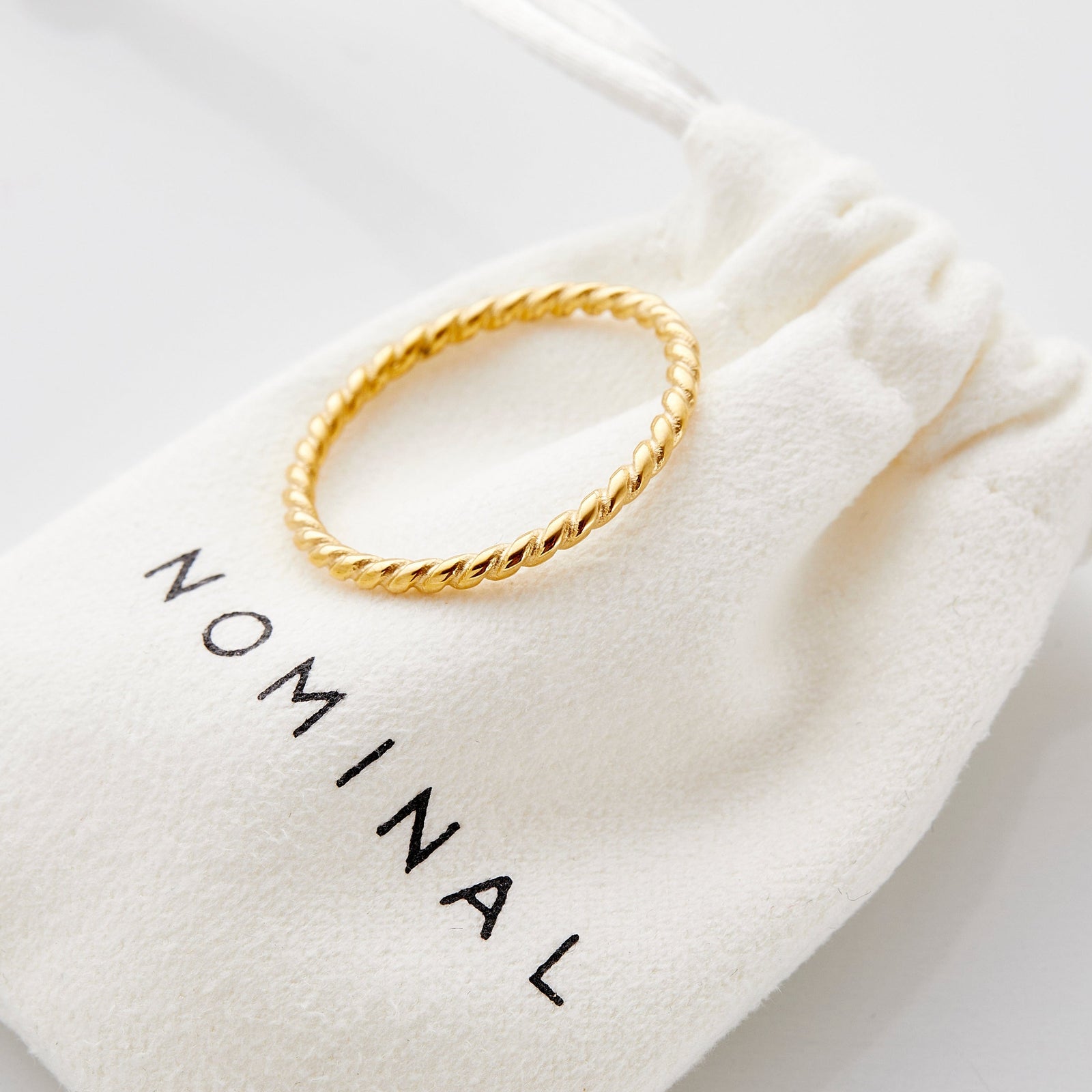 Gold twist ring resting on a white suede pouch with "NOMINAL" text printed in black.