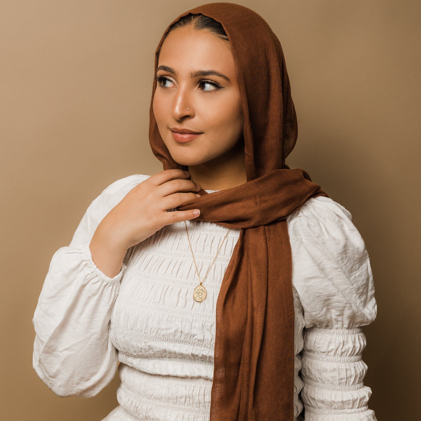Photo of the model looking away from the camera while touching their brown hijab. The model is wearing the gold Guidance Calligraphy Necklace.