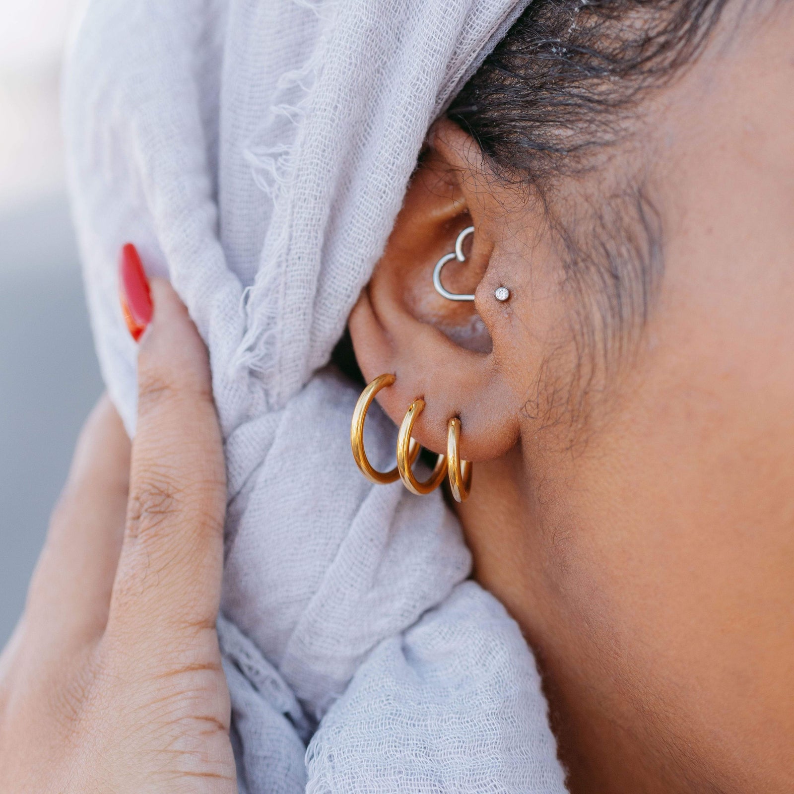 Woman wearing three gold hoop earrings in varying sizes on one ear, styled with a textured light grey hijab and visible cartilage piercings.
