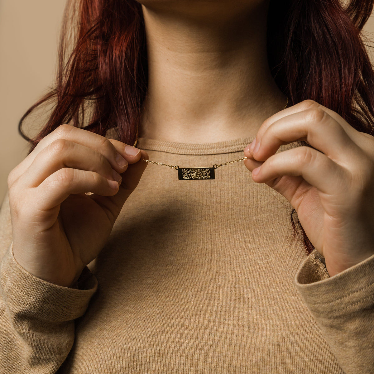Close up of the gold Shahada Bar Necklace as the model lifts it by its chain.